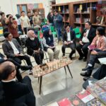 Group of professionals seated in a circle inside a bookstore, discussing with notebooks and phones while photographers document the event.