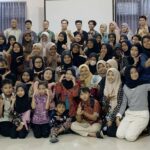 Group photo of students and adults at an indoor event, smiling and posing, with a 'Remaja Geblek Bersastra' banner on the right.