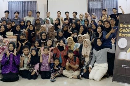Group photo of students and adults at an indoor event, smiling and posing, with a 'Remaja Geblek Bersastra' banner on the right.