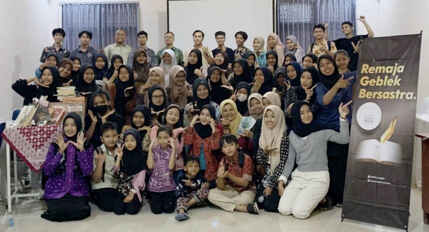 Group photo of students and adults at an indoor event, smiling and posing, with a 'Remaja Geblek Bersastra' banner on the right.