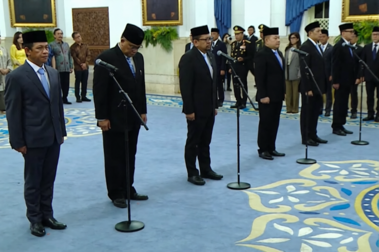 Group of men in suits and traditional caps bowing at mic stands during an official ceremony in a marble-filled room with ornate decor.