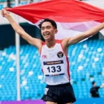 Male athlete finishes a race in a stadium, arms raised, holding the red-and-white Indonesian flag over his head (bib 133).