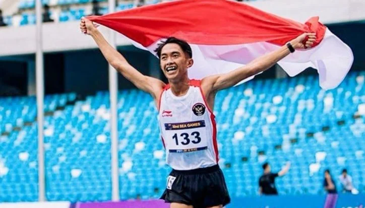 Male athlete finishes a race in a stadium, arms raised, holding the red-and-white Indonesian flag over his head (bib 133).