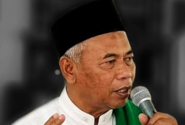 Man wearing a white shirt, black cap, and green scarf speaks into a handheld microphone.