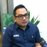 Man with black hair and glasses speaks into a handheld microphone during an indoor interview, with greenery in the background.