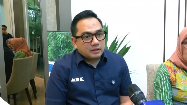 Man with black hair and glasses speaks into a handheld microphone during an indoor interview, with greenery in the background.