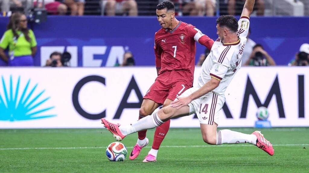 Portugal player wearing red with number 7 controls the ball as an opponent in white slides in a challenge on a green soccer pitch during a match extension of the leg to win the ball. If needed: the image captures an intense moment on the field with a ball near the player's foot and photographers in the background.