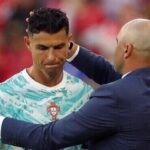 Soccer player in a Portugal jersey being congratulated by a bald coach in a navy suit on the field.