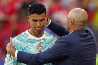 Soccer player in a Portugal jersey being congratulated by a bald coach in a navy suit on the field.
