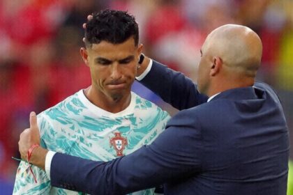 Soccer player in a Portugal jersey being congratulated by a bald coach in a navy suit on the field.