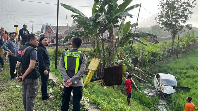 Police officers in bright vests stand by a canal as bystanders observe a rural scene with a child near the water and a tilted metal drum.