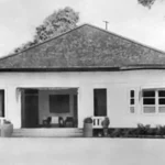 Single-story house with a wide front porch and steps, flanked by trees and a low front wall.