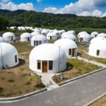 Cluster of white dome-shaped houses with wooden doors in a landscaped village, a person walks along a paved road in the foreground under a blue sky.
