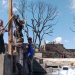 Group of villagers erecting a wooden beam with long poles on a concrete platform, under a blue sky with burnt trees in the background.
