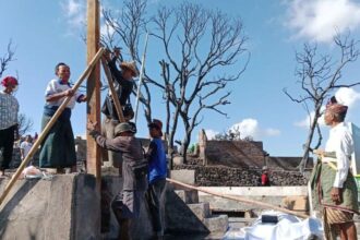 Group of villagers erecting a wooden beam with long poles on a concrete platform, under a blue sky with burnt trees in the background.