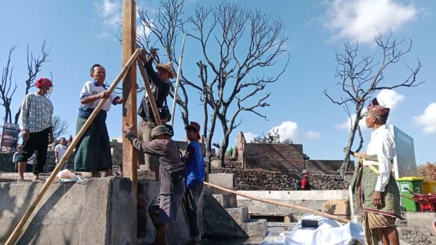 Group of villagers erecting a wooden beam with long poles on a concrete platform, under a blue sky with burnt trees in the background.