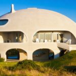 Dome-shaped modern house with arched openings and a carport beneath, on a grassy hill with a driveway and patio seating outside.
