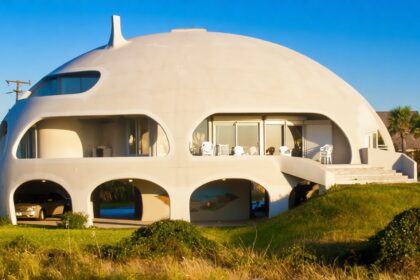 Dome-shaped modern house with arched openings and a carport beneath, on a grassy hill with a driveway and patio seating outside.