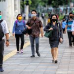 People walking on a city sidewalk while wearing face masks; diverse group in business and casual attire carry bags and backpacks.