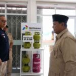 Officials in uniform and civilian attire stand around a shelf displaying Pertamina gas cylinders during a formal event at a facility.