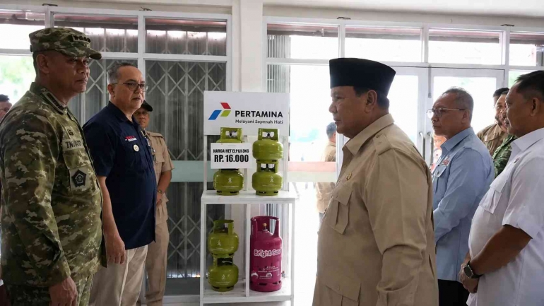 Officials in uniform and civilian attire stand around a shelf displaying Pertamina gas cylinders during a formal event at a facility.