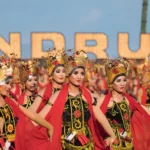 Group of women in ornate traditional costumes dancing in a festival parade, wearing gold headdresses and red capes against a blue sky background.