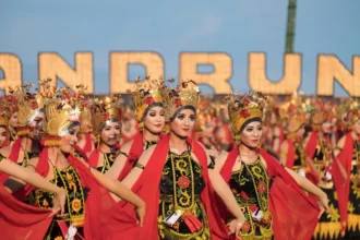 Group of women in ornate traditional costumes dancing in a festival parade, wearing gold headdresses and red capes against a blue sky background.
