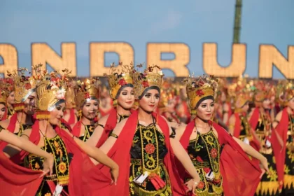 Group of women in ornate traditional costumes dancing in a festival parade, wearing gold headdresses and red capes against a blue sky background.