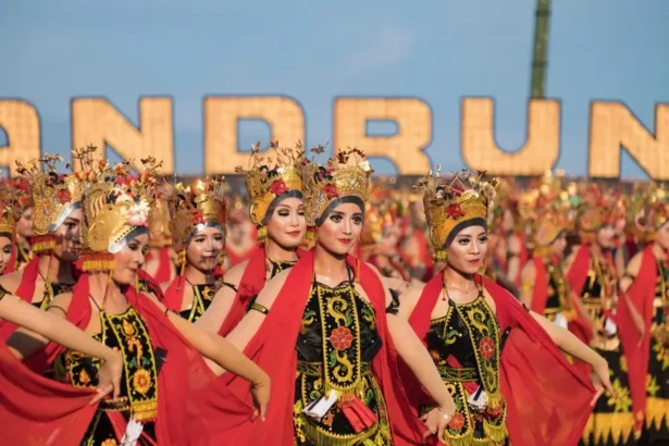 Group of women in ornate traditional costumes dancing in a festival parade, wearing gold headdresses and red capes against a blue sky background.