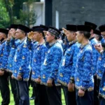 Group of people in blue batik uniforms standing in a line outdoors, many saluting during a formal ceremony.