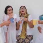 Four women in white outfits laugh together, holding mugs of tea and coffee against a light background.