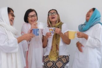 Four women in white outfits laugh together, holding mugs of tea and coffee against a light background.