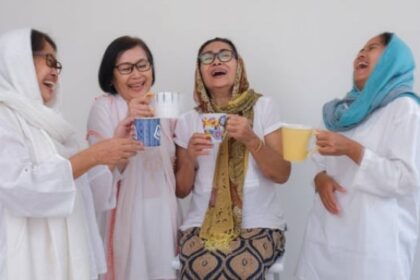 Four women in white outfits laugh together, holding mugs of tea and coffee against a light background.