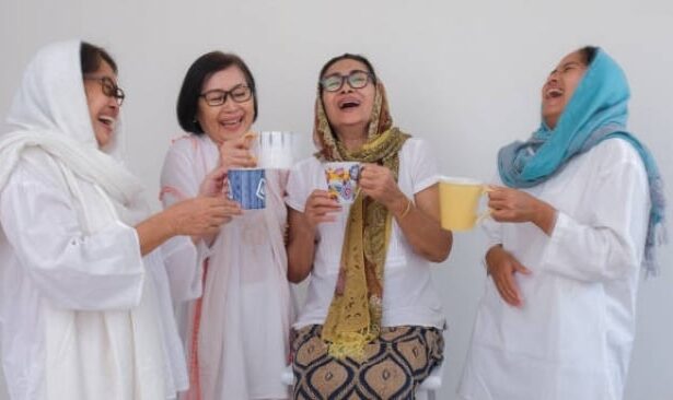 Four women in white outfits laugh together, holding mugs of tea and coffee against a light background.