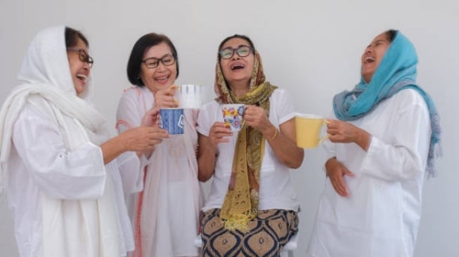 Four women in white outfits laugh together, holding mugs of tea and coffee against a light background.