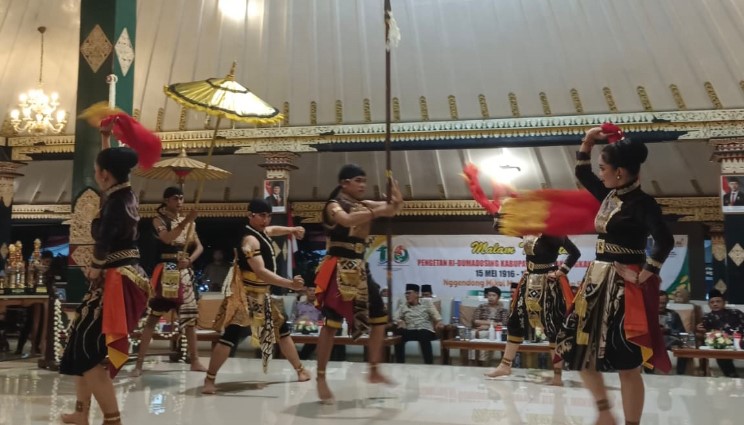 saXZA - Mabur.co Group of Indonesian dancers in colorful costumes performing a traditional dance with fans and parasols indoors, on a polished stage.