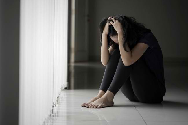 Woman sitting on the floor by a bright window, head in her hands, appearing distressed or overwhelmed.