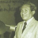 Smiling man in a light suit and tie stands at a podium during a formal event at a conference or ceremony.