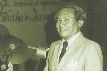 Smiling man in a light suit and tie stands at a podium during a formal event at a conference or ceremony.