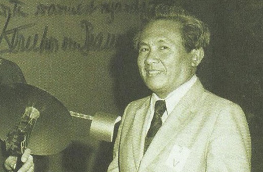 Smiling man in a light suit and tie stands at a podium during a formal event at a conference or ceremony.