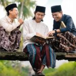 Three men in traditional Malay attire sit on a wooden beam outdoors, reading a book and laughing together.
