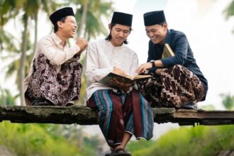 Three men in traditional Malay attire sit on a wooden beam outdoors, reading a book and laughing together.