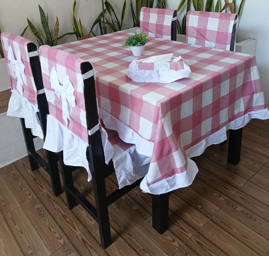 Dining table set with pink-and-white checkered tablecloth and matching chair covers with bows, in a plants-adorned indoor space.