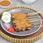 Satay skewers with peanut sauce on a plate, garnished with cucumber slices, onions, and chopped red chili peppers.