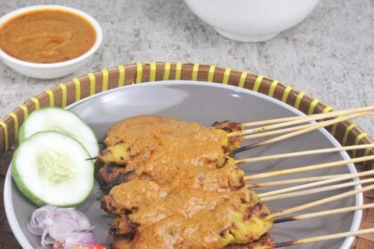 Satay skewers with peanut sauce on a plate, garnished with cucumber slices, onions, and chopped red chili peppers.