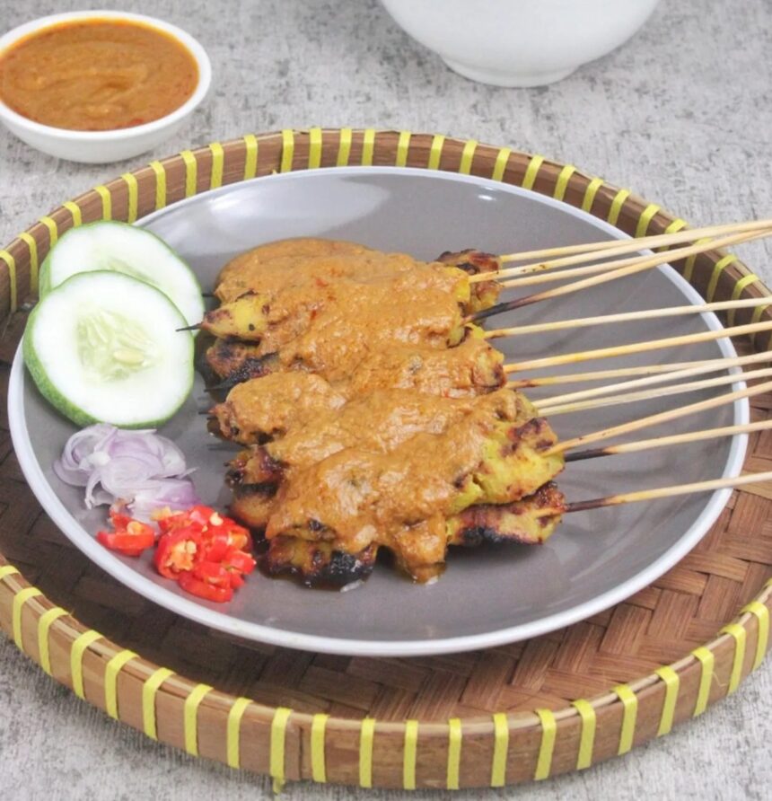 Satay skewers with peanut sauce on a plate, garnished with cucumber slices, onions, and chopped red chili peppers.