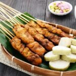 Basket of fried meat skewers on a banana leaf, with white yam chunks and a dipping sauce nearby in a wicker tray on a dark wooden table.