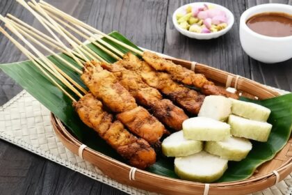 Basket of fried meat skewers on a banana leaf, with white yam chunks and a dipping sauce nearby in a wicker tray on a dark wooden table.