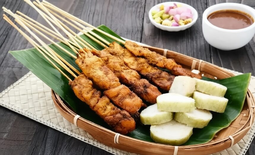 Basket of fried meat skewers on a banana leaf, with white yam chunks and a dipping sauce nearby in a wicker tray on a dark wooden table.
