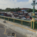 View of a city bridge with green and yellow railing, round bollards, and a river below, with densely built houses nearby.